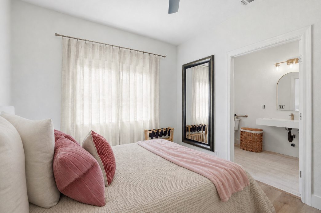 A private bedroom in cream and sage — framed landscape art, a lit table lamp, and garden windows beside the bed