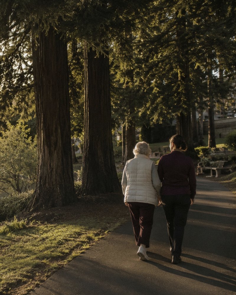 Two women walking a redwood-lined path beside the water in late-afternoon light
